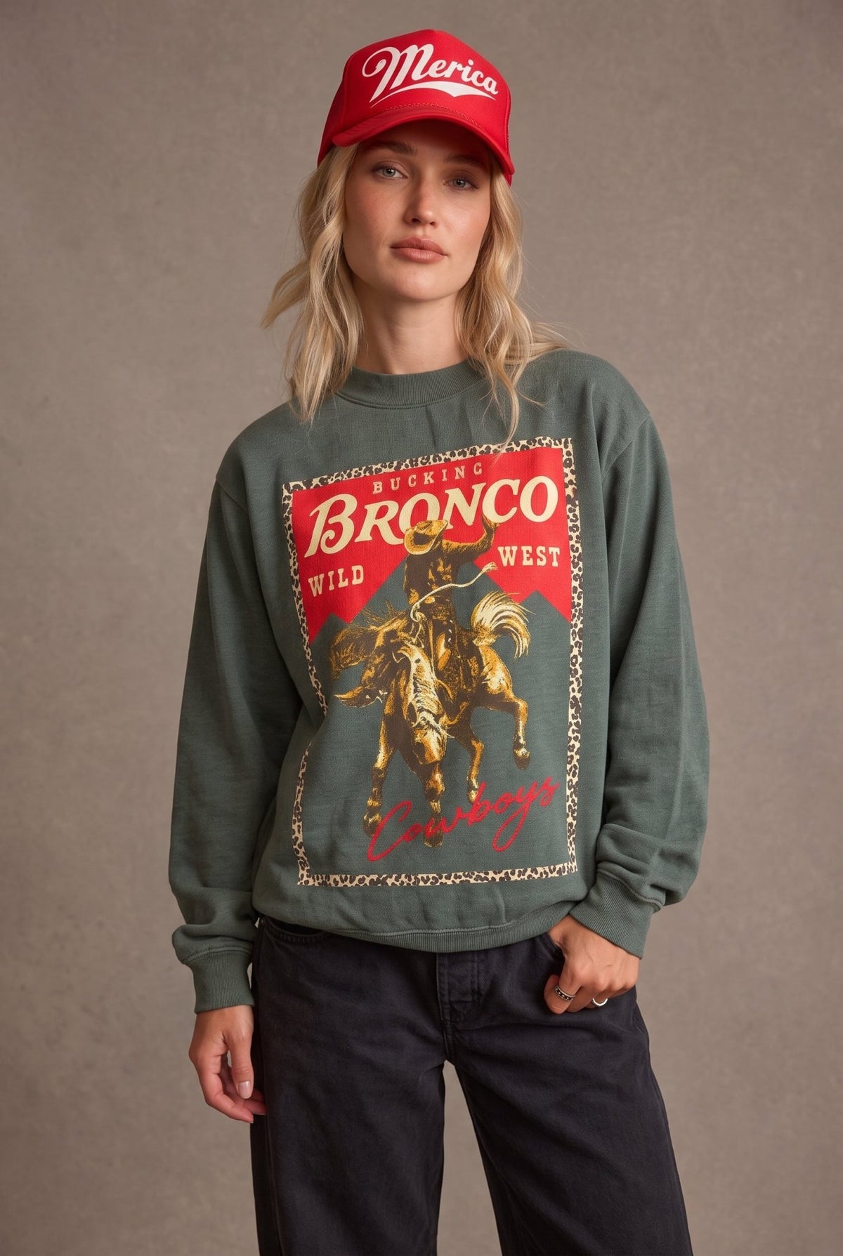 Women's vintage washed sage green western graphic crewneck sweatshirt featuring an oversized, relaxed silhouette, classic round neckline, dropped shoulders, long sleeves, and ribbed cuffs. The front showcases a bold retro rodeo-inspired graphic with a bucking bronco horse and cowboy rider in distressed golden yellow and brown tones, set against a vibrant red background with a leopard print border. Featuring the words "Bucking Bronco," "Wild West," and "Cowboys" in vintage western typography, this cozy pullo