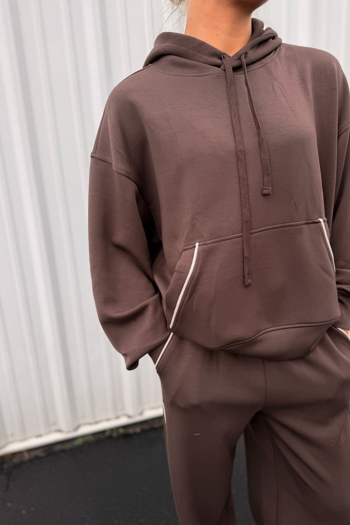 Women’s chocolate brown pullover hoodie featuring a relaxed oversized fit, drawstring hood, dropped shoulders, long sleeves, front kangaroo pocket, and contrast ivory piping detail along the pocket and sides. Coordinating chocolate brown wide-leg sweatpants are available separately for a matching lounge set.