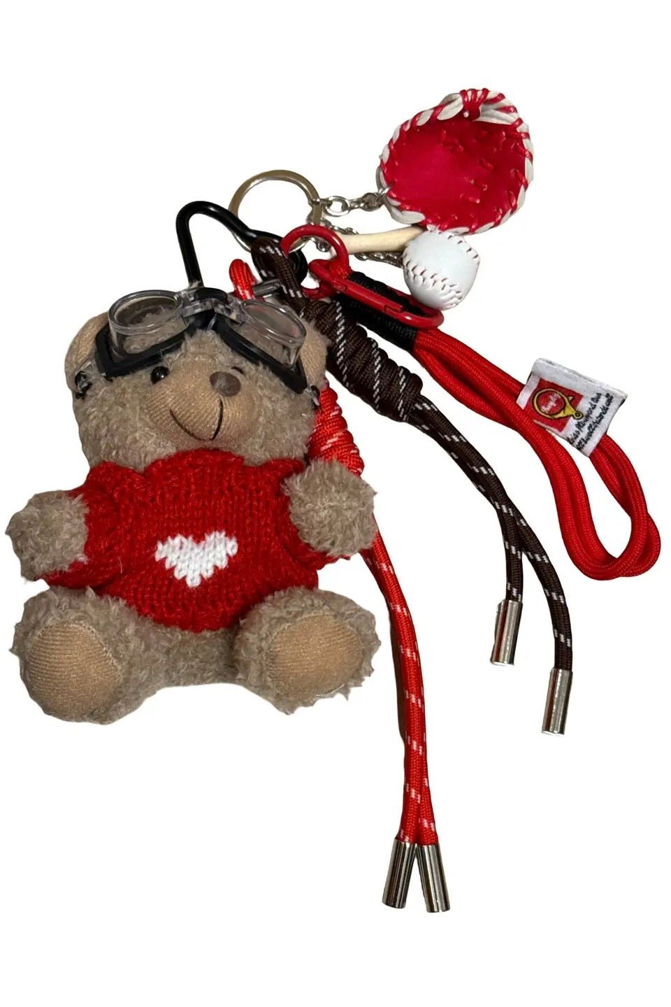 Teddy bear keychain wearing red sweater with white heart, black goggles, with miniature baseball, red baseball mitt, wooden bat, and red and brown braided rope tassels, on a white background.