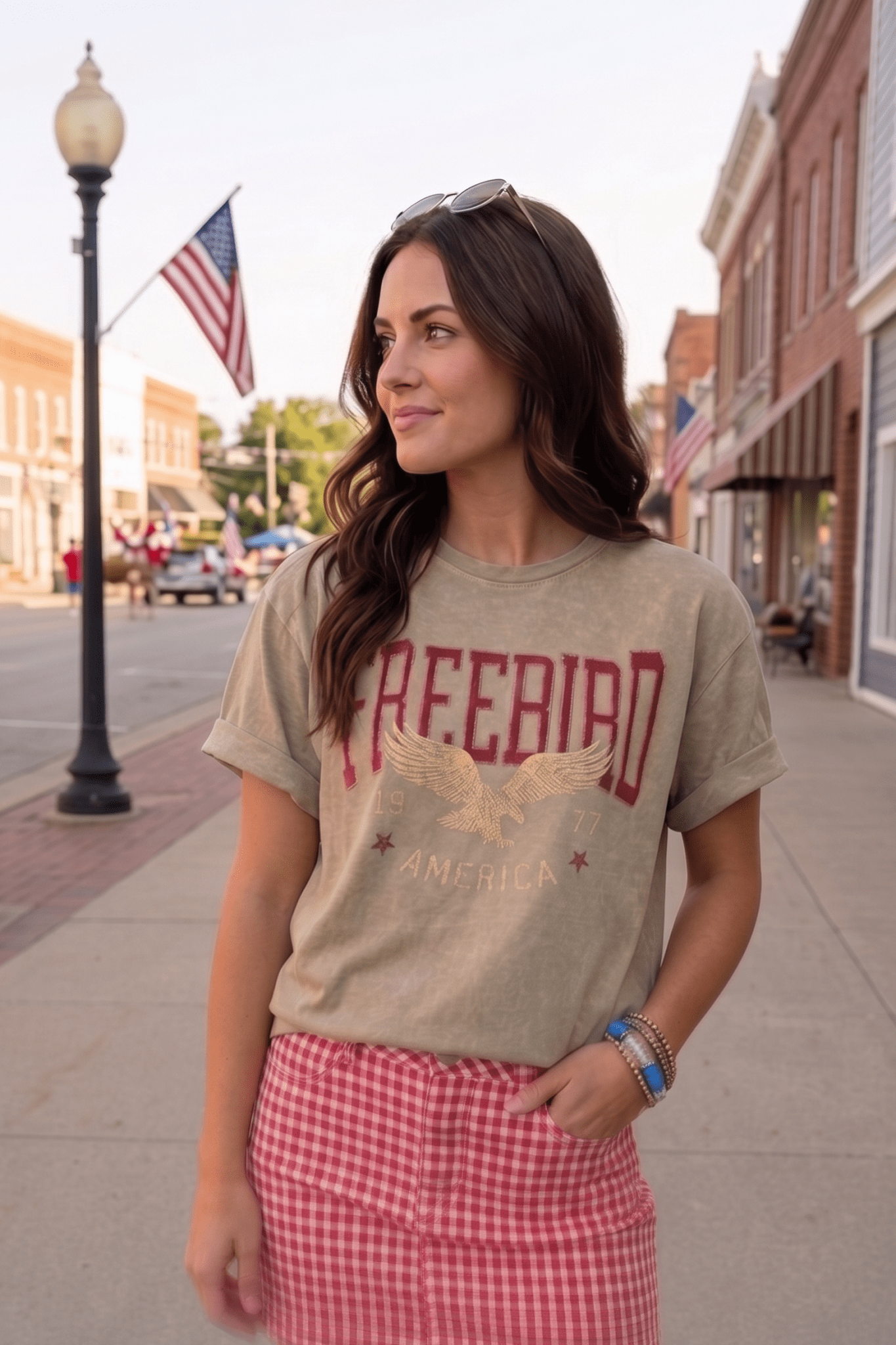 Women's Free Bird America 1977 graphic tee featuring a vintage eagle graphic and oversized fit. Styled with a red gingham skirt for a country concert outfit, rodeo outfit, Fourth of July look, western festival outfit, and Americana summer fashion.