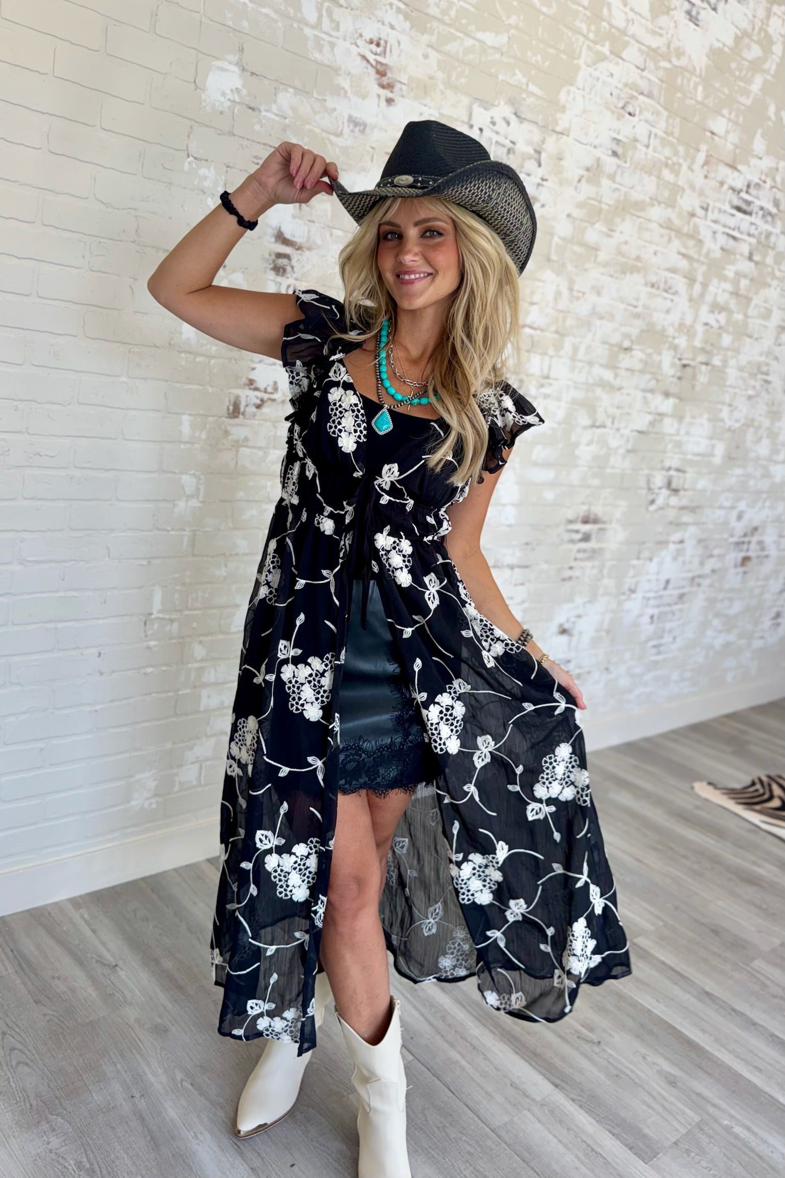 Woman wearing a black sheer embroidered chiffon duster with flutter sleeves, sweetheart neckline, tie-front detail, floral embroidery, denim shorts, and white western boots.