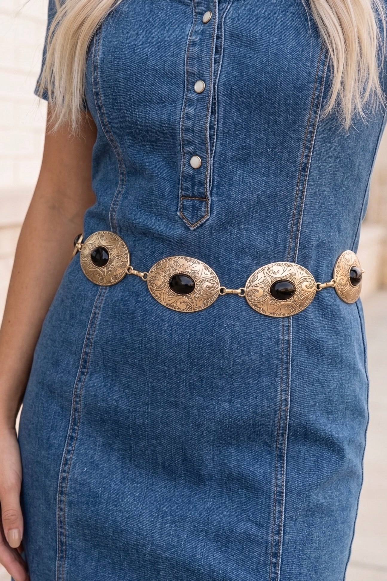 Western gold-tone chain concho belt with engraved oval conchos and black stone accents styled around the waist over a cream dress.