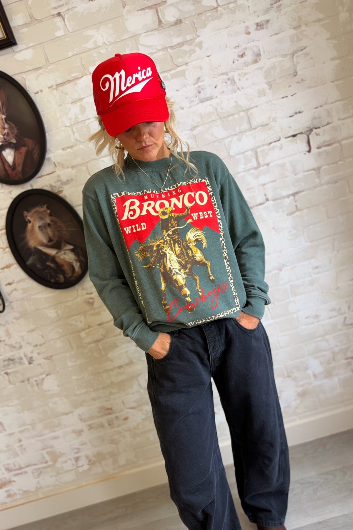 Women's vintage washed sage green western graphic crewneck sweatshirt featuring an oversized, relaxed silhouette, classic round neckline, dropped shoulders, long sleeves, and ribbed cuffs. The front showcases a bold retro rodeo-inspired graphic with a bucking bronco horse and cowboy rider in distressed golden yellow and brown tones, set against a vibrant red background with a leopard print border. Featuring the words "Bucking Bronco," "Wild West," and "Cowboys" in vintage western typography, this cozy pullo