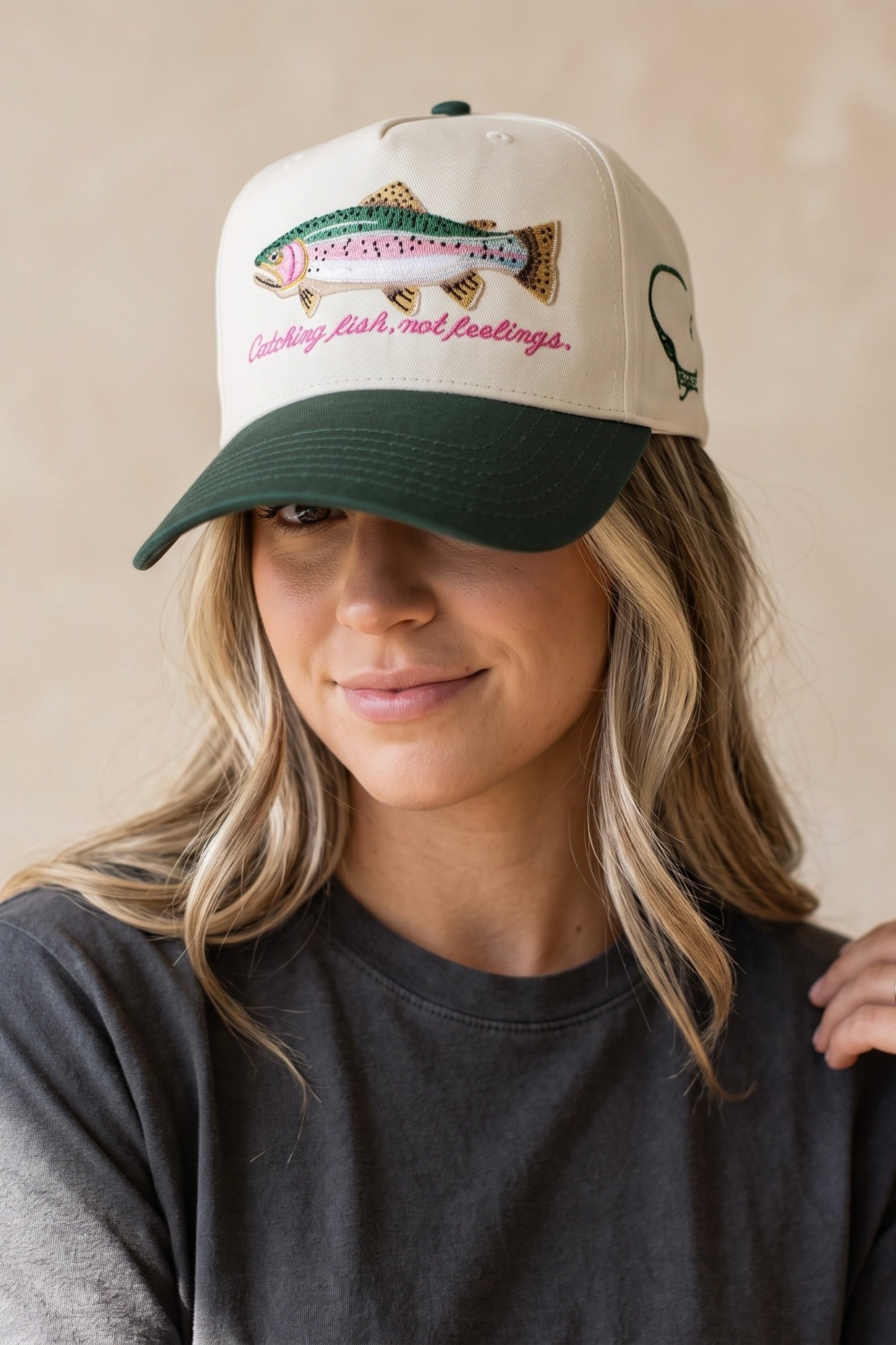 Cream and forest green trucker hat featuring an embroidered rainbow trout with the phrase "Catching Fish, Not Feelings" and an adjustable mesh back.