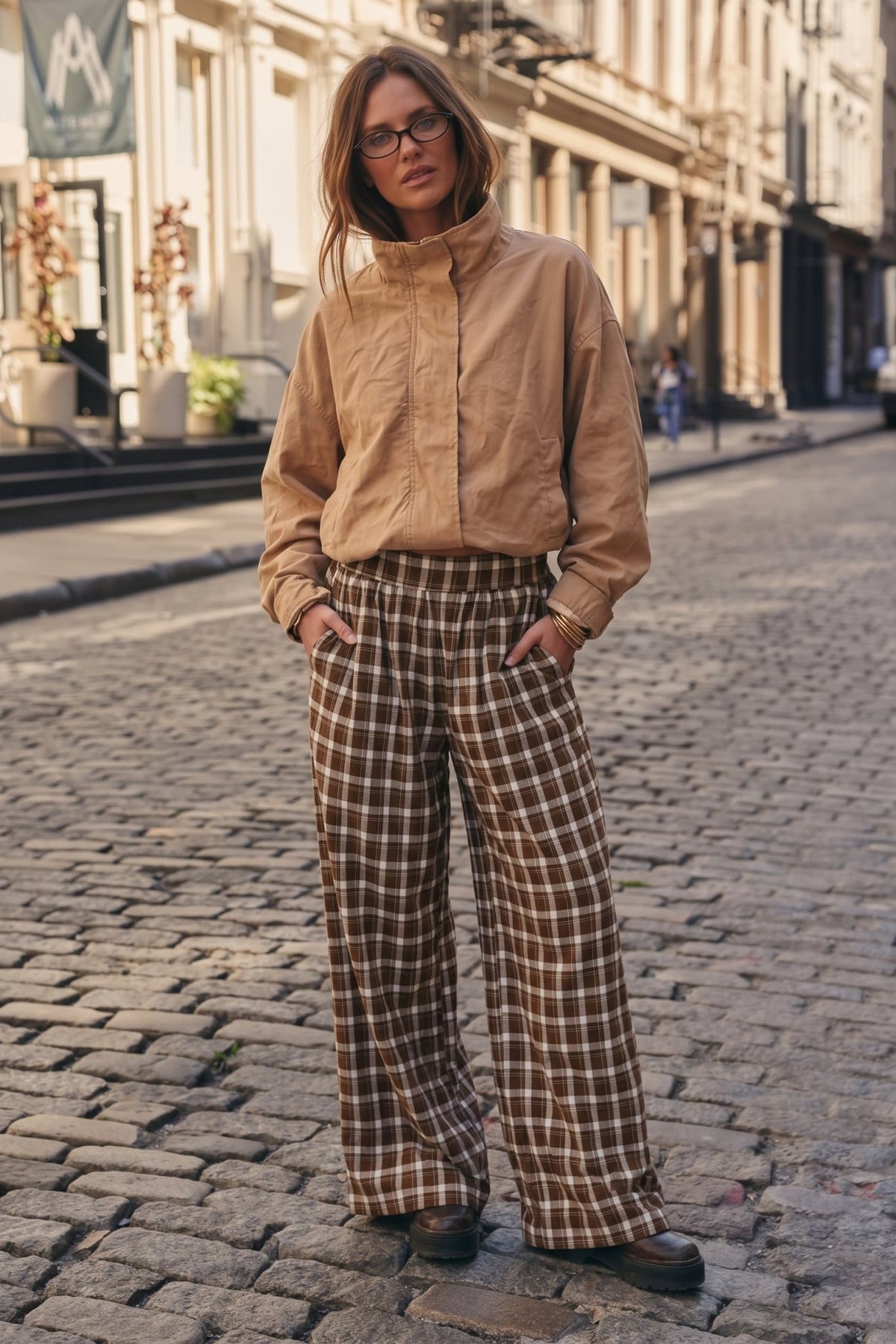 Woman wearing brown, cream, and white plaid wide-leg pants featuring an oversized, flowy silhouette, relaxed fit, and side pockets, styled with a cropped tan jacket.